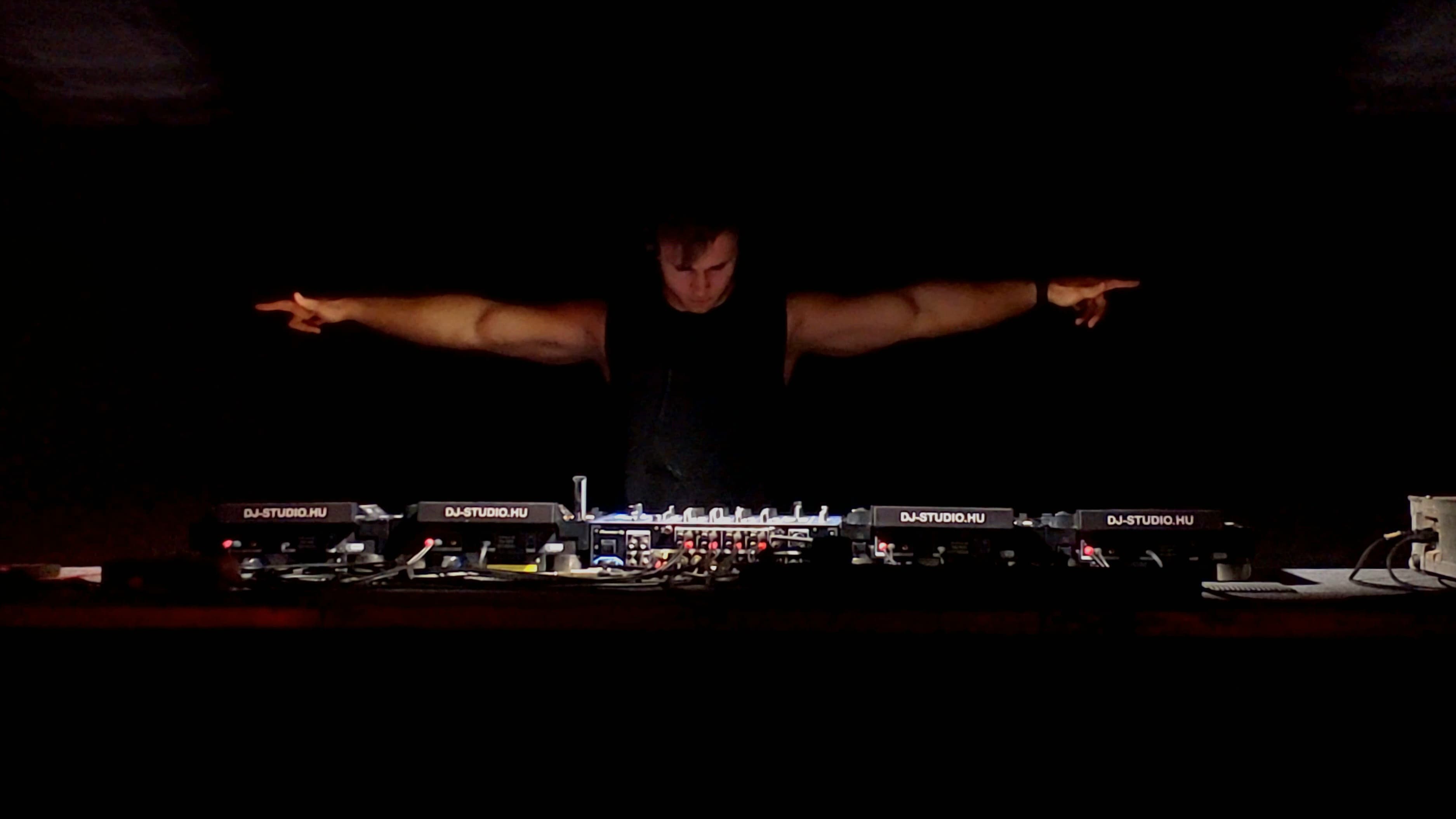 FRIXX behind a row of DJ decks with both arms extended in a dark venue.