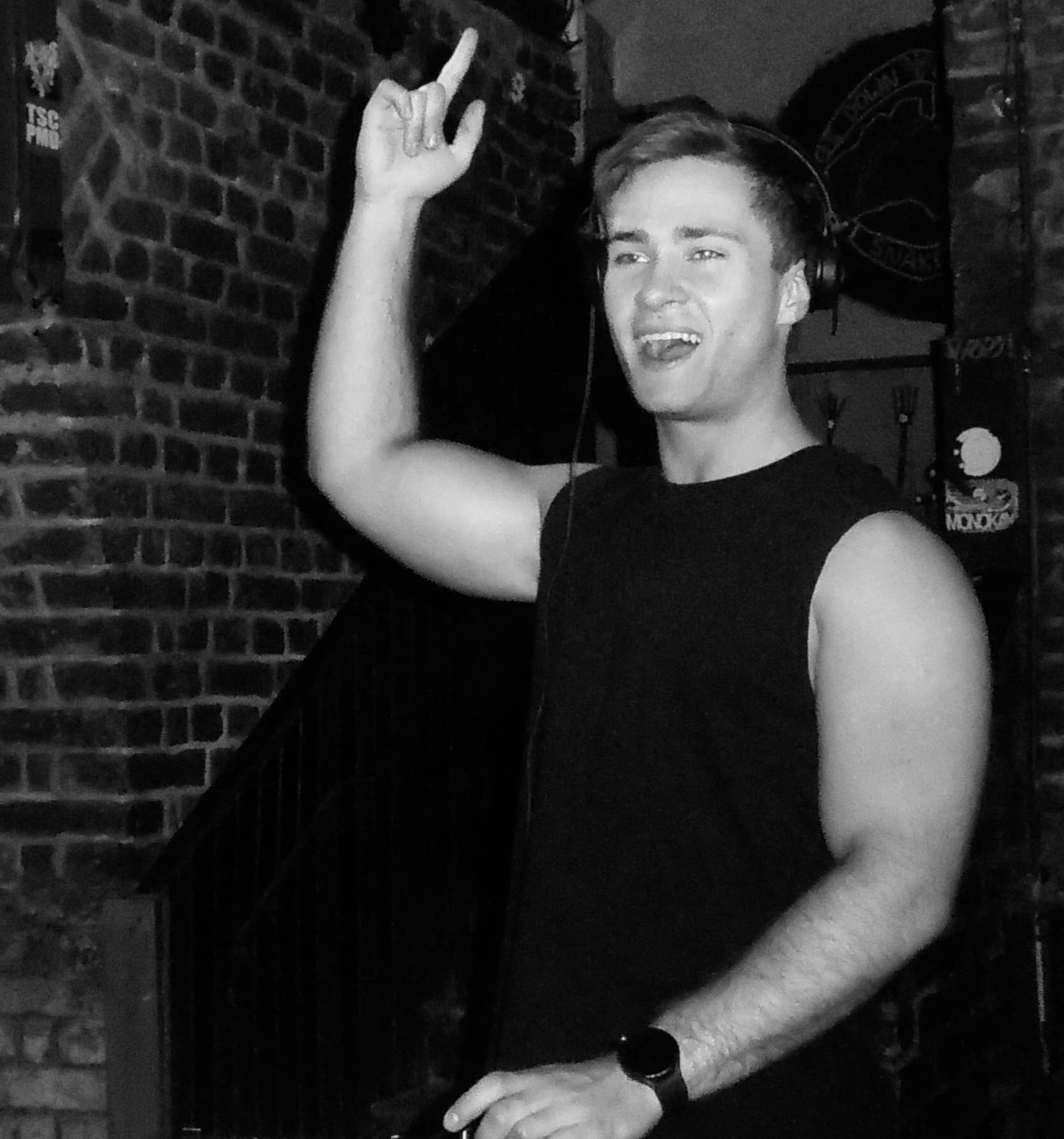 Black-and-white photo of FRIXX smiling behind the decks with one hand raised.